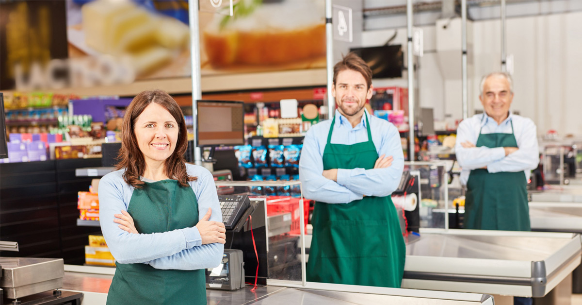 Tres trabajadores de un supermercado posan sonrientes junto a la línea de cajas. 